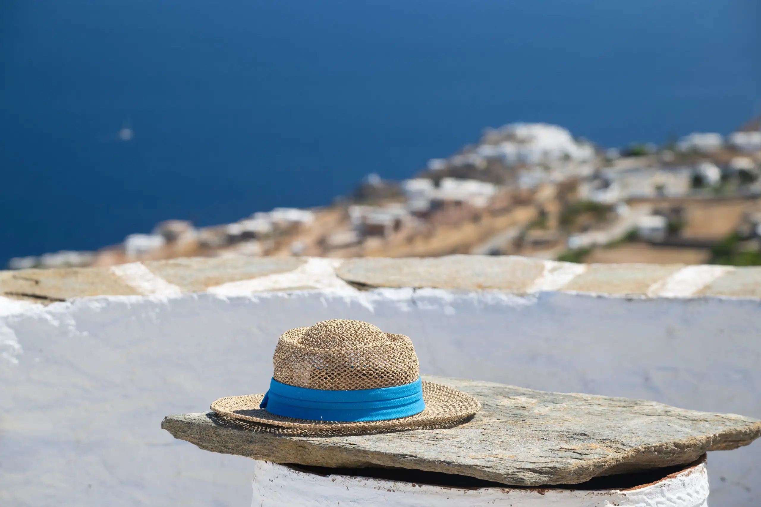 Ψάθινο καπέλο σε ασβεστωμένο τοιχάκι, με χωριό της Σίφνου και τη θάλασσα στο βάθος